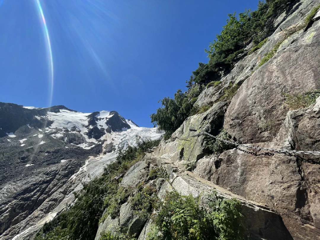A chain bolted along a cliffside trail, glaciated peaks across the valley