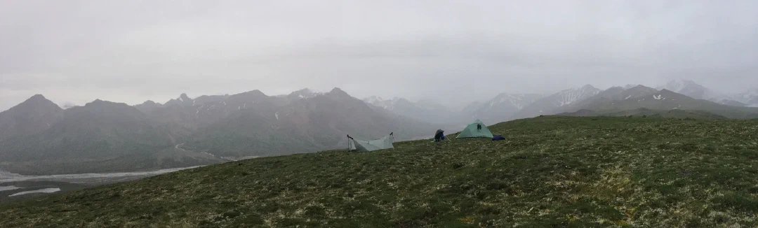 Tarp and tent shelters pitched on a tundra crest above a misty sweep of grey ridgelines and glacial melt.
