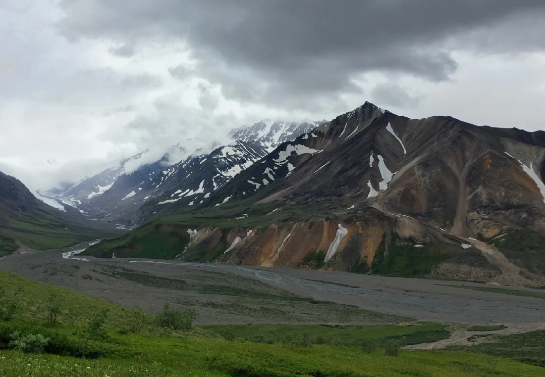 Cloud spilling over a snow-and-rust mountain face above a glacier-fed river bar.