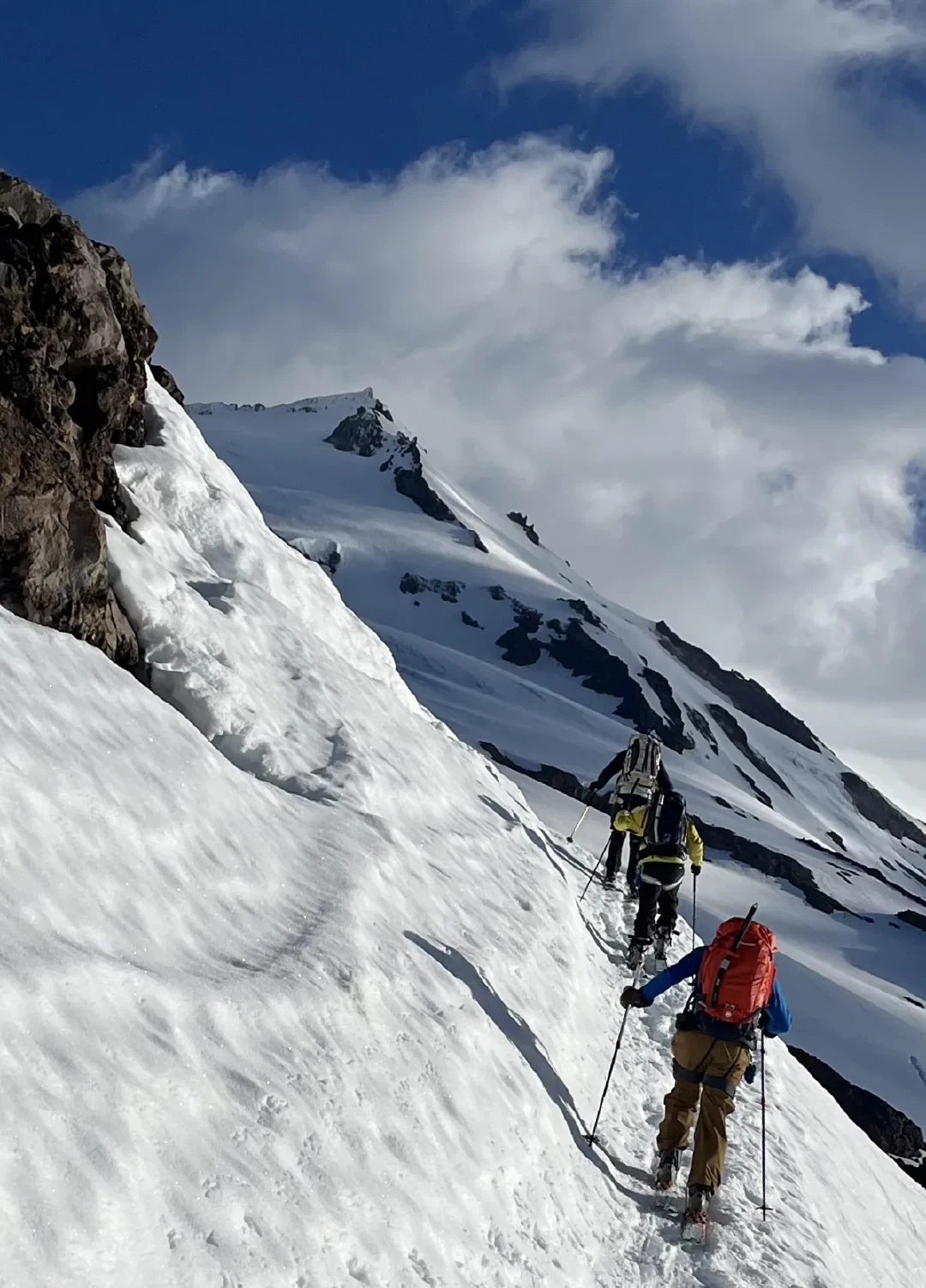 Three skiers skinning up beneath a rocky spur, the false summit of Sherman Peak beyond.