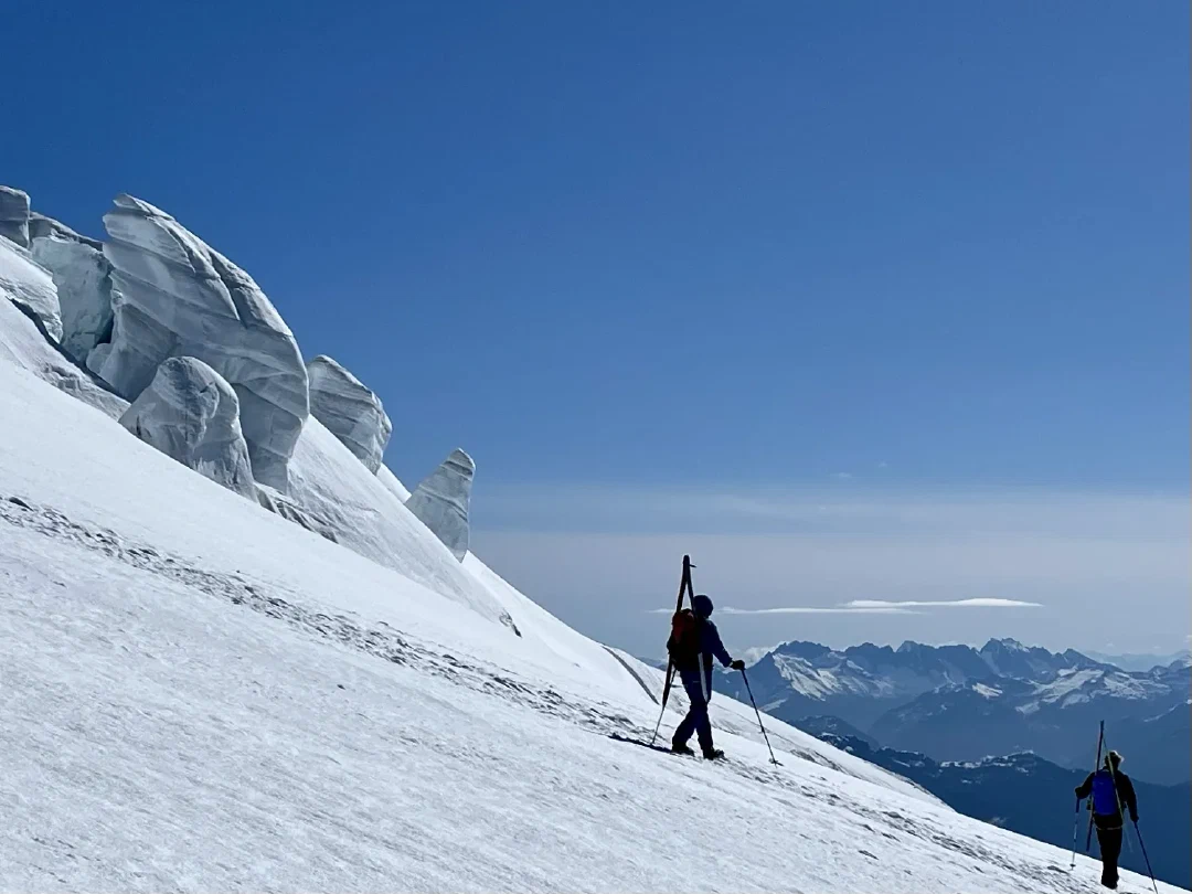 Two skiers traverse Squak Glacier beneath tilted seracs, skis on packs.