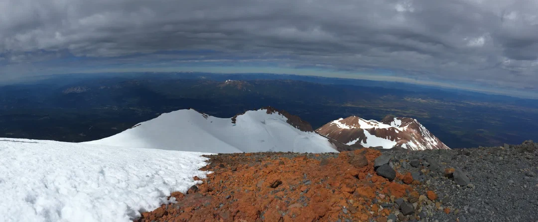 Panorama from the summit: snowfields and red volcanic rock dropping away to hazy valleys under layered clouds