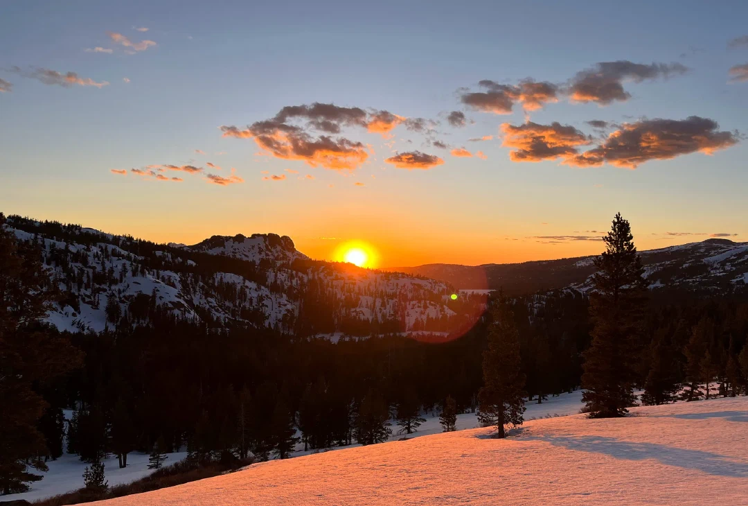 Sun dropping behind a rocky ridge, snowfields in the foreground glowing pink.