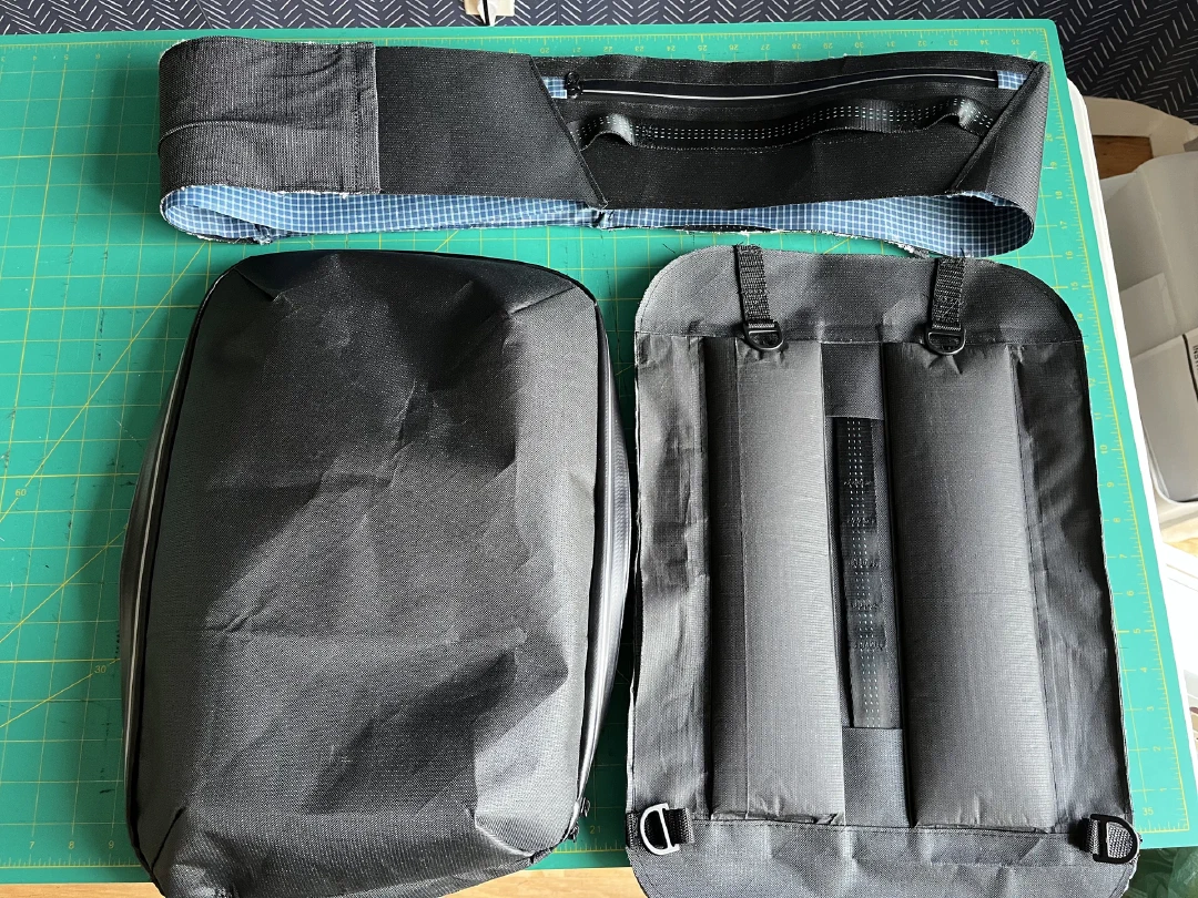 Three finished subassemblies side by side: zippered top panel, shell body, and padded harness panel with a retracting sheathed elastic grab handle.