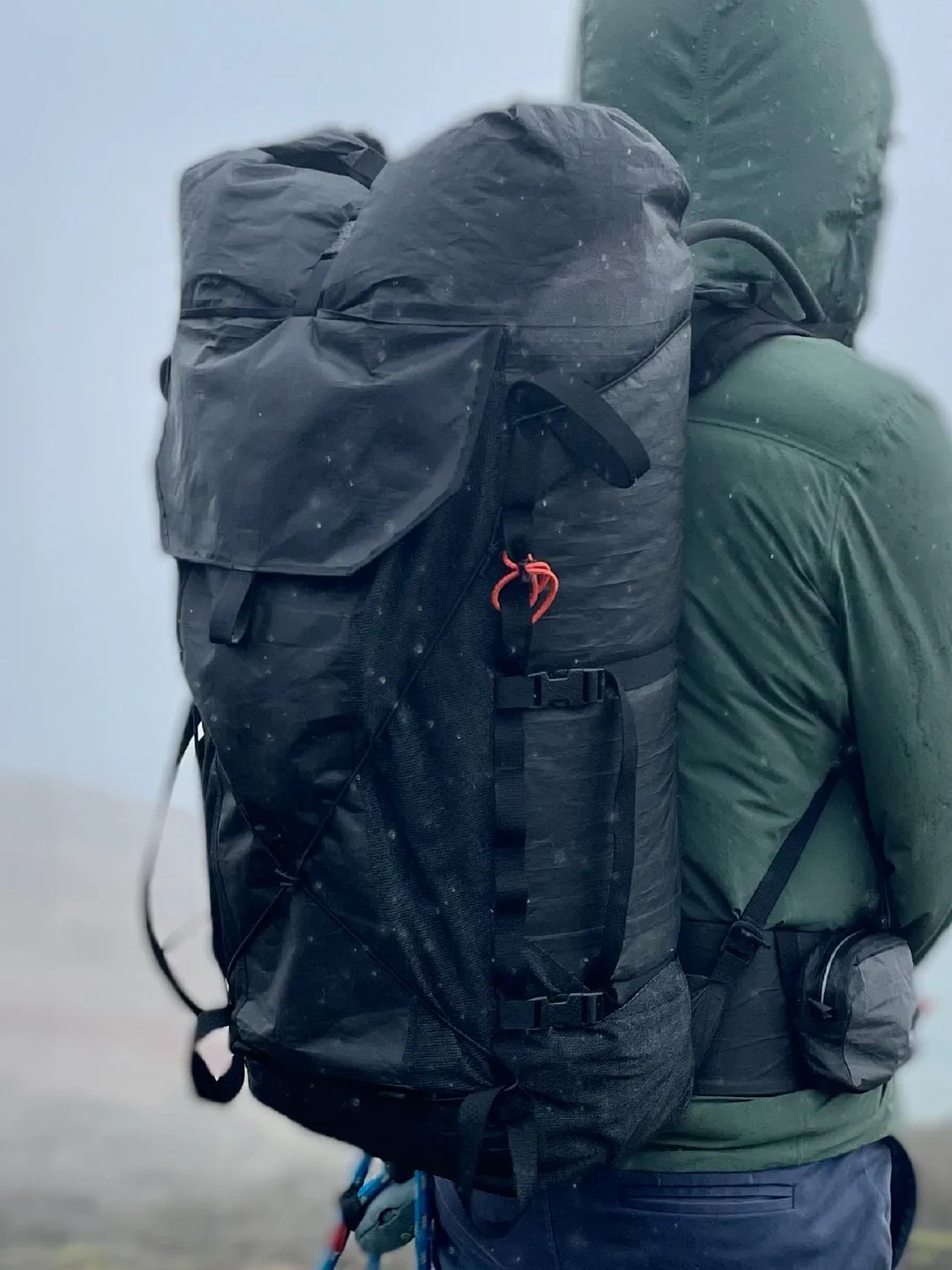 Loaded pack worn from behind in coastal rain and fog, rolltop cinched, with optional hipbelt pouch and hydration hose routed through shoulder port.